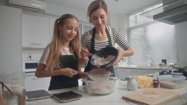 Medium shot of Caucasian mother teaching daughter to make dough for cookies sifting flour in bowl