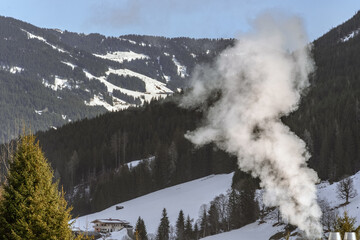 A serene winter landscape featuring a snow-covered mountain, dense forest, and a building emitting steam, illustrating the concept of eco-friendly living amidst nature