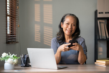 Mature asian woman using a smartphone at her desk with a laptop in a home office. Remote work and...