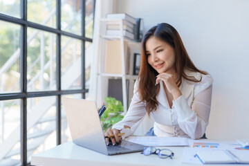 Professional asian businesswoman with a confident smile in a bright office setting with a laptop and documents.