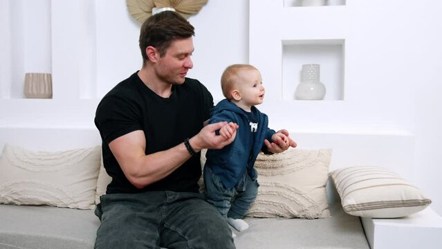 Caucasian father with an infant baby sitting on the sofa. Cute little child are dancing.