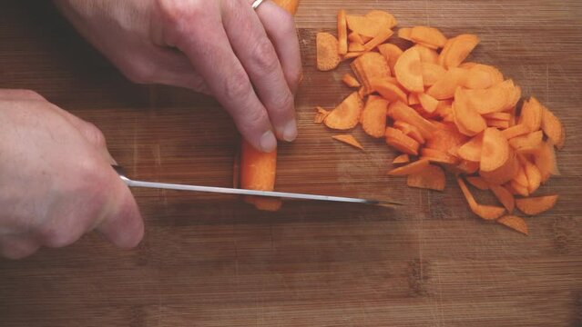 Preparing vegetable salad. Man chopping carrot with knife on wooden cutting board. Top View Male Chef Knife Cut Vegetables. Slice carrots for healthy vegan recipe. Close-up slow motion knife cutting