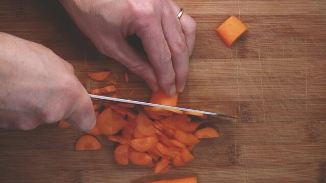 Top view of man slices orange carrots with a metal knife vegan food. Top down video knife slicing carrots on a wooden board. Close-up slow motion of knife cutting a orange carrots on the wooden board.