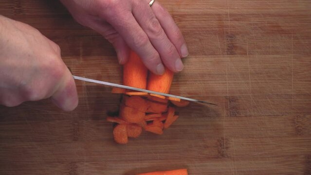 Close up slow motion of knife cutting a orange carrots on the wooden board.Top view of man slices orange carrots with a metal knife vegan food. Top down video knife slicing carrots on a wooden board.