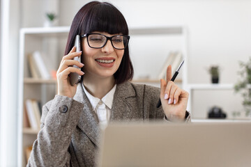 Experienced employee coming up with idea of improving company productivity in workplace. Joyful lady in formal wear speaking on smartphone while reading report from digital screen.