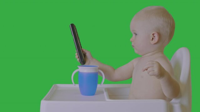 Infant in highchair with remote control, green screen background, early technology interaction. Baby hold remote, focus attention, potential chroma key visual effects. Entertainment education concept.