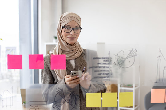 Portrait of beautiful muslim woman standing in front of glass board and typing on smartphone. Female office worker in hijab and stylish suit using digital phone in company.
