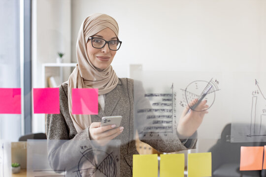 Portrait of beautiful muslim woman standing in front of glass board and typing on smartphone. Female office worker in hijab and stylish suit using digital phone in company.