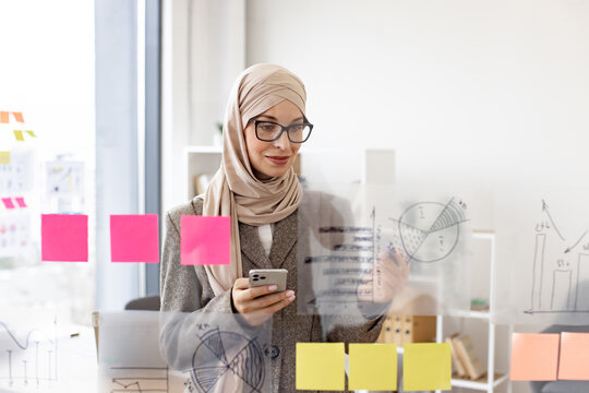 Pensive arab businesswoman looking on phone while analyzing growth of charts on glass board at modern office. Concept of business strategy and digital marketing.