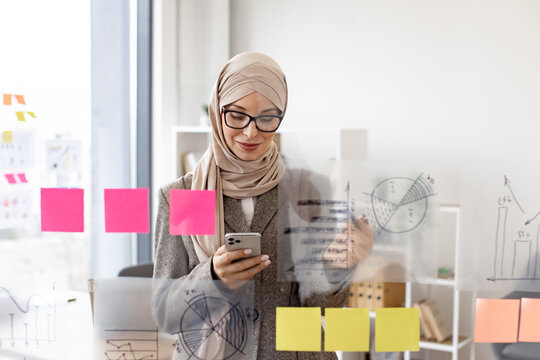 Pensive arab businesswoman looking on phone while analyzing growth of charts on glass board at modern office. Concept of business strategy and digital marketing. - Powered by Adobe