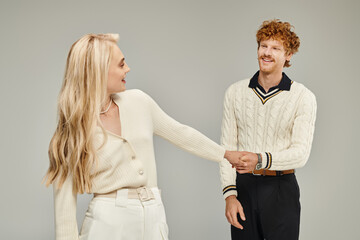 cheerful blonde woman and stylish redhead man holding hand on grey, old money style concept