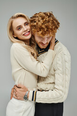 smiling and stylish blonde woman hugging young redhead man on grey, fashionable couple relationship
