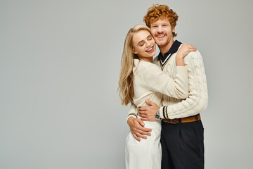 happy and trendy young couple in stylish casual attire embracing on grey, classic timeless fashion