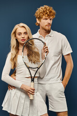 trendy blonde woman with tennis racquet looking at camera near redhead man in white attire on blue