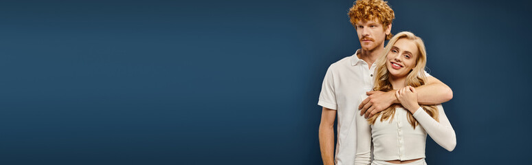 young redhead man embracing joyful blonde woman in white stylish attire on blue backdrop, banner