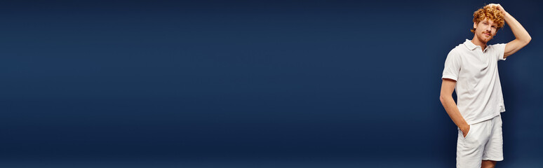 young redhead man in white stylish clothes posing with wand in pocket on blue, horizontal banner