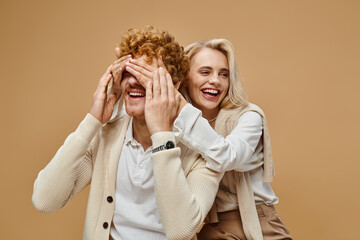 playful blonde woman covering eyes of stylish redhead man on beige, old money style fashion