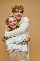 happy redhead man in eyeglasses embracing trendy blonde woman on beige, old money style couple