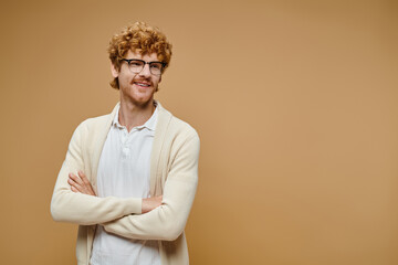 happy redhead man in eyeglasses and stylish clothes posing with folded hands looking away on beige
