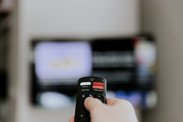 Hand holding remote control pointing to TV. Person watching smart television indoors. Blurred...