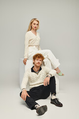 trendy redhead man looking at camera near blonde woman sitting on white cube on grey, classic style