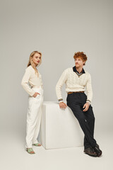 blonde woman and redhead man in trendy casual attire posing near white cube on grey backdrop