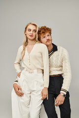 dreamy redhead man hugging trendy blonde woman while sitting on white cube on grey, classic fashion