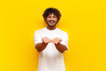 indian man in white t-shirt holds empty hands in front of him and begs on a yellow isolated...