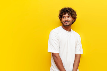 modest indian man in white t-shirt is shy on a yellow isolated background, an insecure guy thinks...