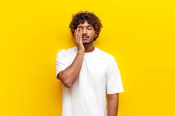 Fotobehang tired indian man in white t-shirt suffering from headache and sad in depression on yellow isolated background, unhappy curly guy rubbing his eyes © Богдан Маліцький