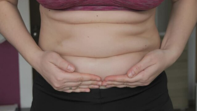 Female belly body positive. Woman touches belly folds in flat slow motion. A small layer of fat on abdomen of woman wearing a sports bra and leggings. Concept of sport, healthy lifestyle or overweight