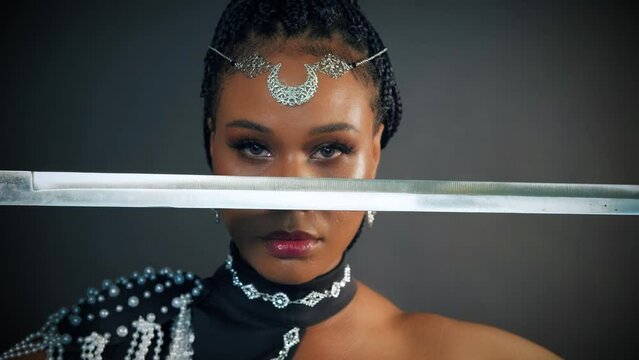 Art portrait african fantasy woman warrior queen holding sword in hands hiding face eyes with sharp blade, sexy mystic image. Beauty lady in black dress girl princess tiara moon, dark gray studio.