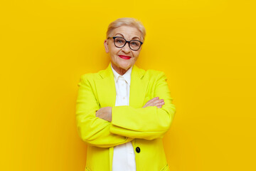 old businesswoman in glasses and business clothes stands with her arms crossed on yellow isolated...