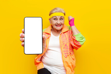 funny old granny in sports colorful clothes wins online and shows blank smartphone screen on yellow...