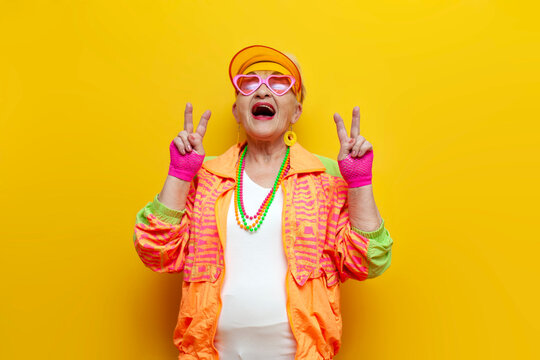 crazy old granny in sports colorful clothes wins and showing peace gesture on yellow isolated background, elderly woman in youth hipster clothes rejoices in victory and luck and screams
