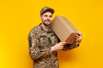 Ukrainian army soldier in pixel camouflage uniform holds a box on a yellow isolated background, Ukrainian military cadet carries and gives humanitarian aid and donation - obrazy, fototapety, plakaty