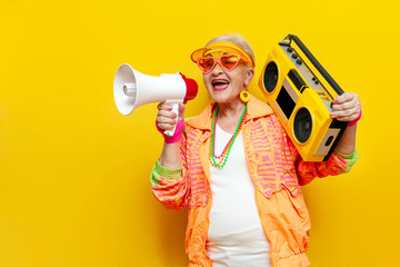 funny crazy granny in hipster clothes listens to music on a tape recorder and announces information into a megaphone on a yellow isolated background, elderly cool woman dancing at a party and shouting