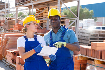 African-american man and European young woman in yellow hardhats talking about documentation in outdoor construction material storage.