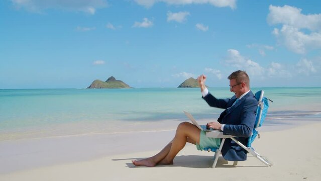 Happy young businessman celebrating victory, clenching fist with excitement, smiling, working online, sitting on the beach with white sand and azure ocean. High quality 4k footage