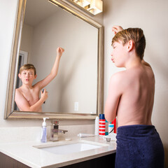 Preteen boy standing at bathroom mirror spraying deodorant under his arm