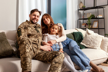 Fototapeta na wymiar Ukrainian army soldier in camouflage uniform returned home to his family, military cadet sits on the sofa with his wife and daughter and smiles, child hugs veterans father