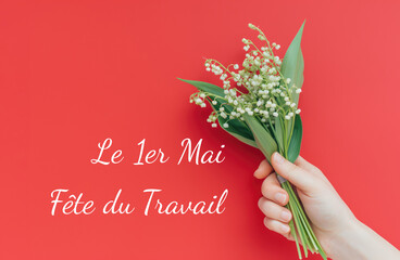 A hand presents a bouquet of Lilies of the Valley against a blue background with text celebrating May Day, Labor Day in France..