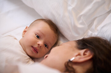Close-up portrait of a cute baby boy looking at camera, lying on the soft white bed sheets close to his loving mother