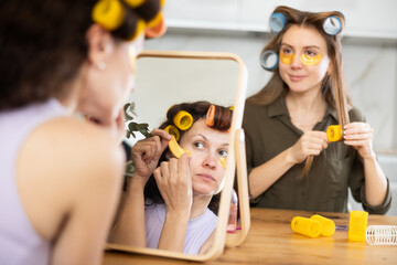 In front of vanity mirror, woman with best friend with hair rollers on heads enjoying skincare...