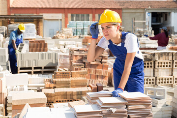 Caucasian young woman in yellow hardhat standing beside tile stack in outdoor outdoor construction material storage.