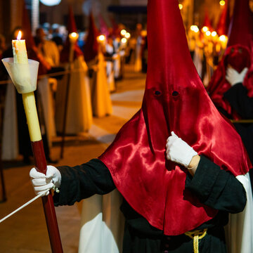 Candle held by unrecognized Member of a brotherhood dressed in tunic and red capirucho in an Holy Week procession at night. Devotee, copy space, nazarene