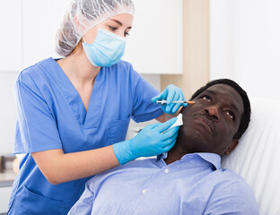 Fototapeta premium Skilled female cosmetologist performing face contouring injections to adult african american man in cosmetology clinic. Male skin care concept