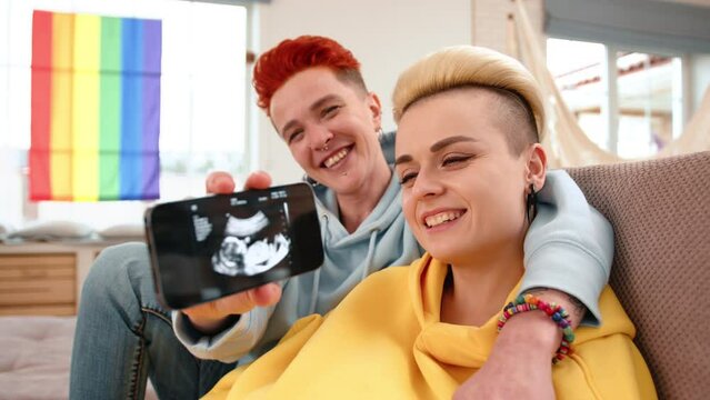 A lesbian couple sits comfortably at home, smiling as they proudly display an ultrasound image on their phone, a colorful LGBT flag in the background. High quality 4k footage