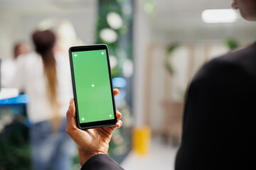 Woman holding mobile phone with green screen in hand while shopping in boutique. African american woman using smartphone with blank chroma key display in department store