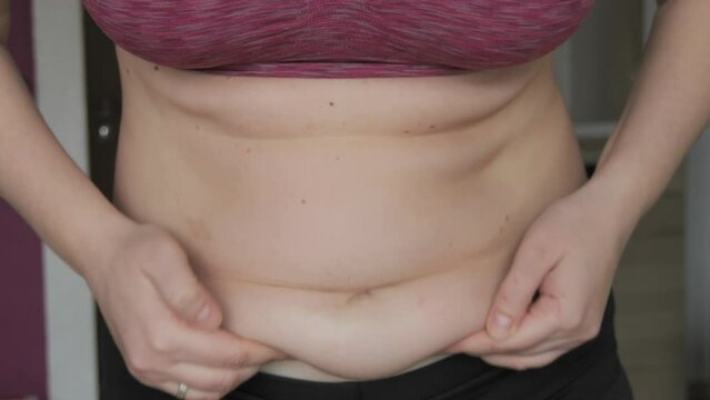 Female belly body positive. Woman touches belly folds in flat slow motion. A small layer of fat on abdomen of woman wearing a sports bra and leggings. Concept of sport, healthy lifestyle or overweight